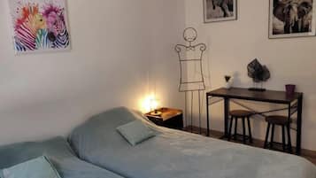 A room with a bed, a desk with two chairs, a small table with a plant, and two framed pictures on the wall.