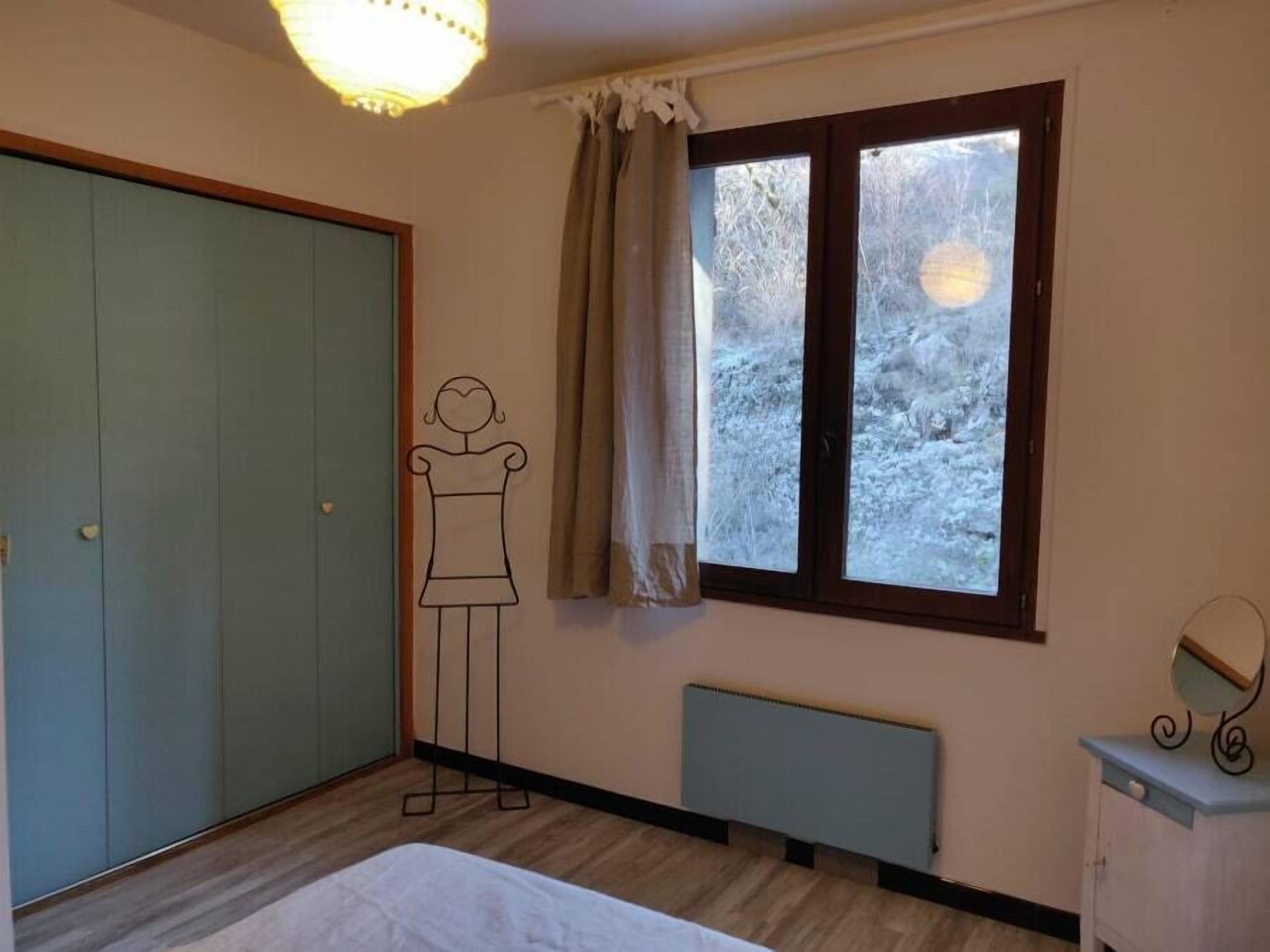 Un dormitorio con cama, armario, radiador, ventana con cortinas y espejo adosado a la pared.