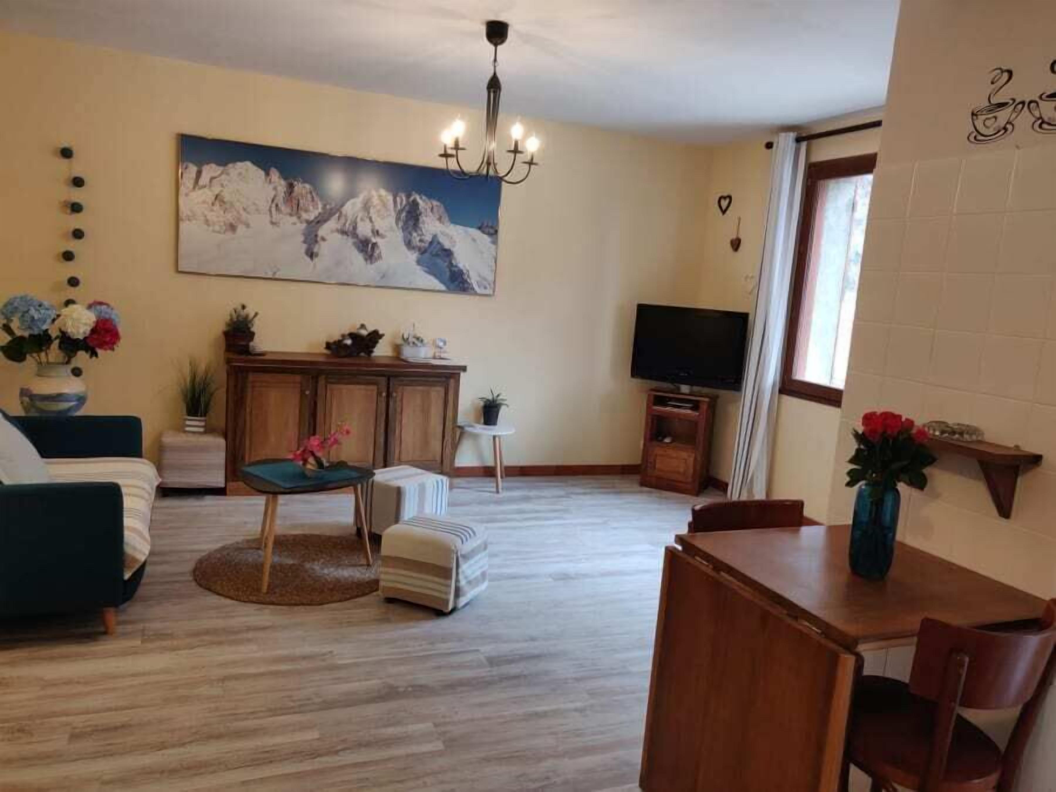 Una sala de estar con un sofá, una mesa de comedor con sillas, un televisor, una araña y un cuadro de montañas en la pared.