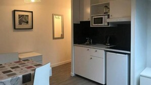 Egy kis méretű konyha fehér szekrényekkel, mikrohullámú sütővel és kávéfőzővel. Van egy étkezőasztal székekkel, és a falon egy kép található keretben.