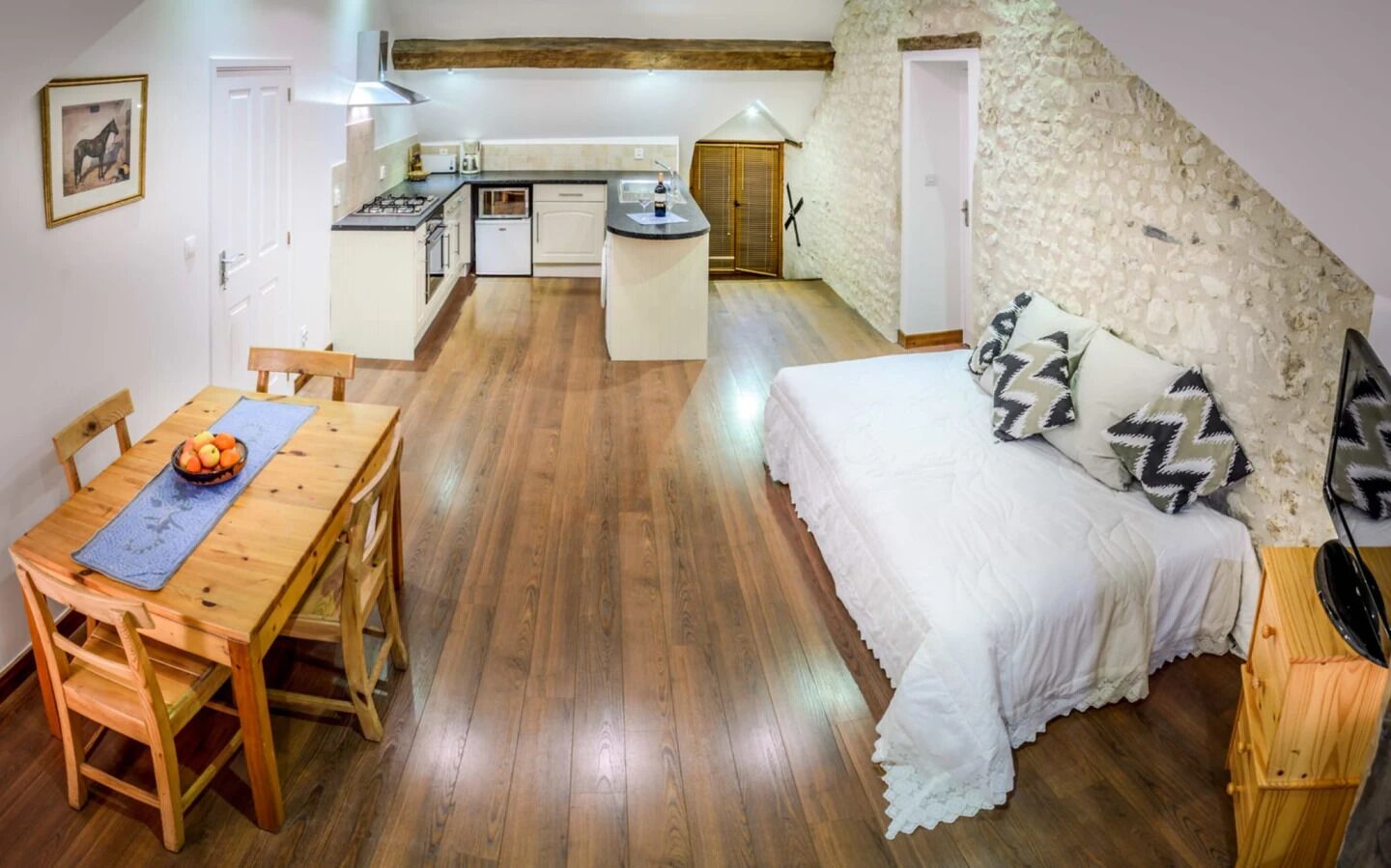 Une pièce chaleureuse avec une table à manger en bois, un lit agrémenté d’oreillers à motifs et un coin cuisine avec un mur en pierre.