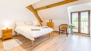 Ein Schlafzimmer mit Holzboden, einem Bett mit weißer Bettdecke, einem Nachttisch aus Holz, einem Stuhl und Blick ins Freie durch eine Tür.