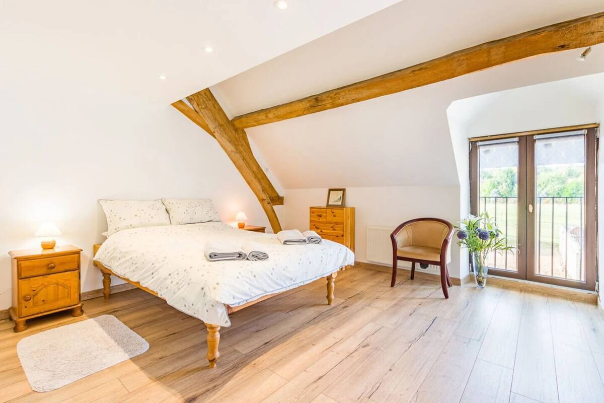 A bedroom with a wooden ceiling, a bed with a white bedspread, a wooden nightstand, a chair, and a view of the outdoors through a door.