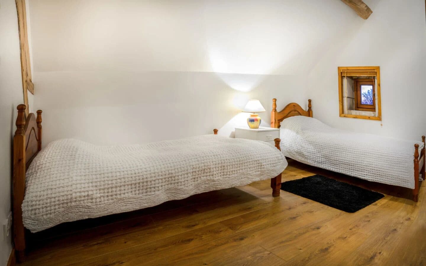 A bedroom with two single beds, wooden headboards, a nightstand with a lamp, and a small window with a wooden frame.