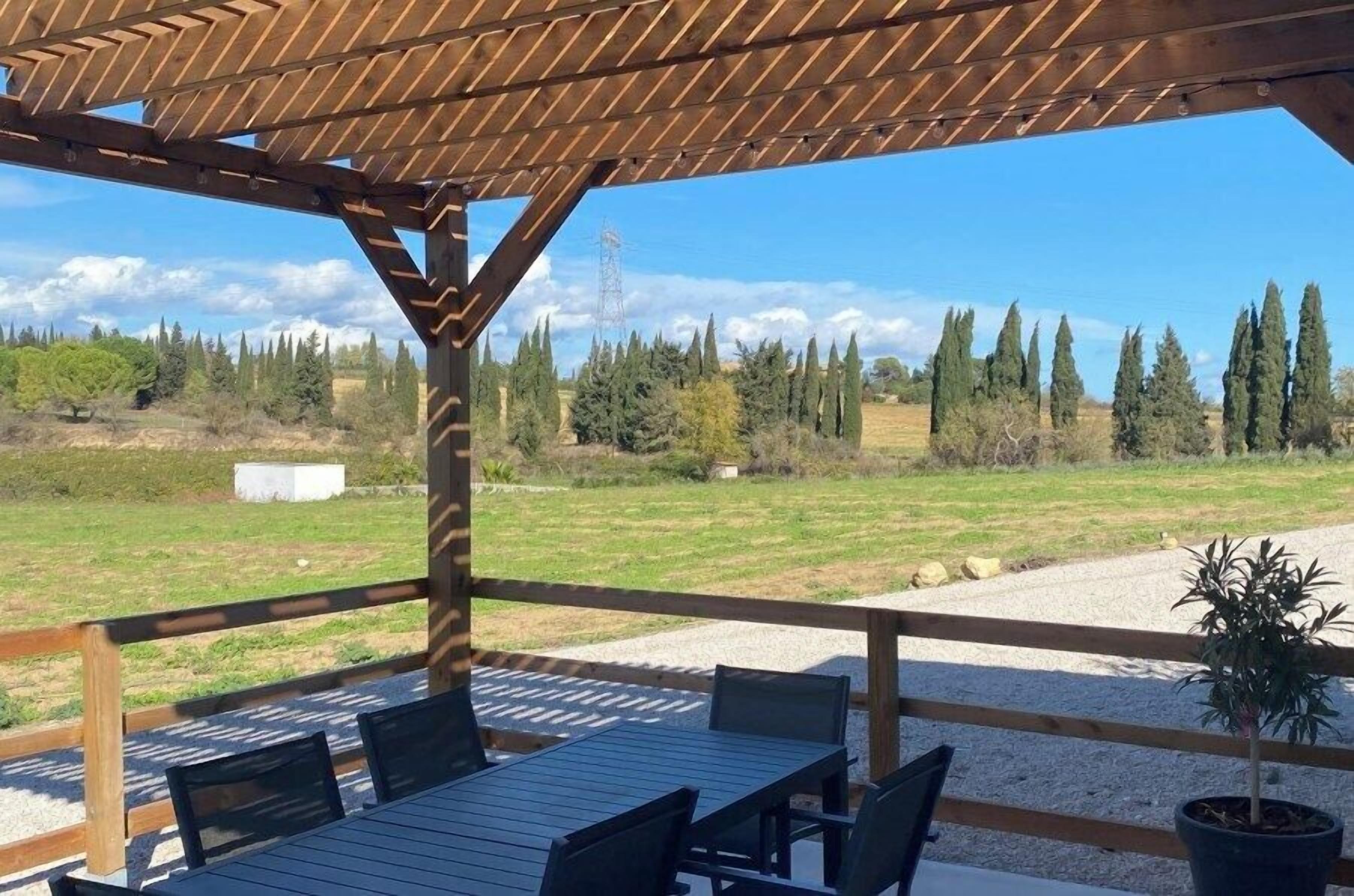 An outdoor wooden pergola with a table and chairs, overlooking a grassy field with trees and a power line in the background.