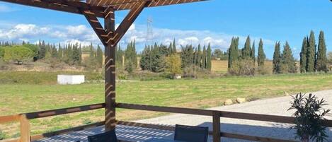 An outdoor wooden pergola with a table and chairs, overlooking a grassy field with trees and a power line in the background.