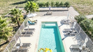 Une piscine extérieure entourée de palmiers et d’une clôture, avec des chaises longues et des parasols.