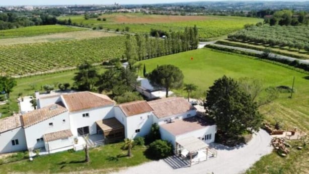 Un bâtiment blanc, doté d’un toit recouvert de tuiles, entouré de champs verdoyants et d’arbres.
