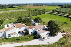 Un bâtiment blanc, doté d’un toit recouvert de tuiles, entouré de champs verdoyants et d’arbres.