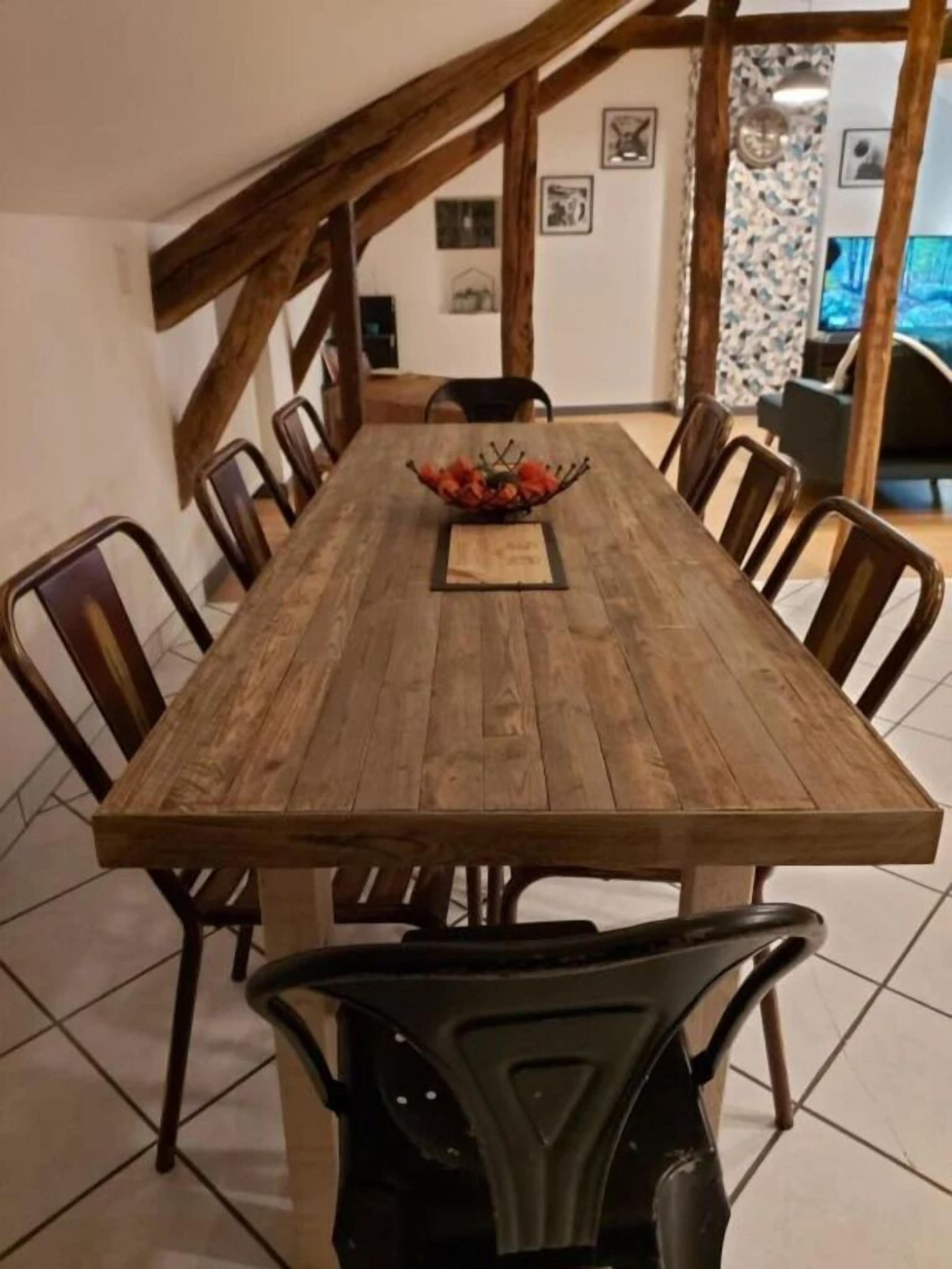 A dining table with a wooden finish, surrounded by six chairs, in a room with exposed wooden beams and tiled flooring.