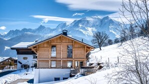 A wooden chalet with a sloped roof, surrounded by snow-covered mountains and a clear sky.