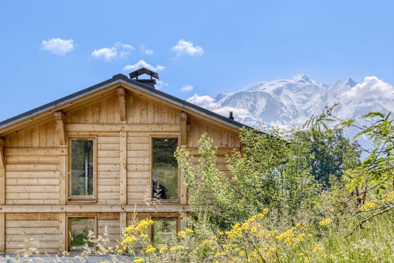 A wooden cabin with a large window and a door, surrounded by wildflowers and trees, with a mountain range in the background.