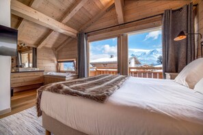 Un dormitorio con una cama grande, vigas de madera en el techo, vistas a montañas y un balcón con baranda de madera.