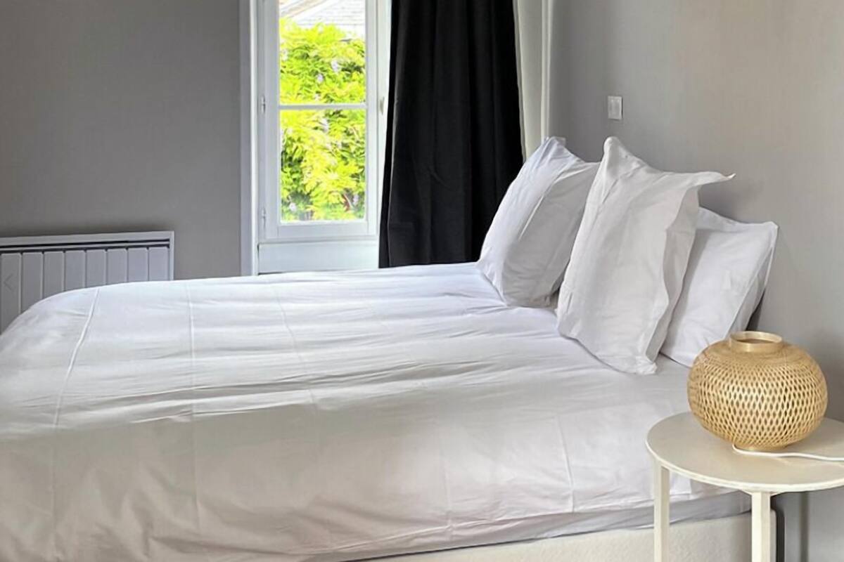 Ein ordentlich bezogenes Bett mit weißen Leinen und Kissen, eine runde gewebte Lampe auf einem kleinen weißen Nachttisch und ein Fenster mit schwarzen Vorhängen, das natürliches Licht hereinlässt.