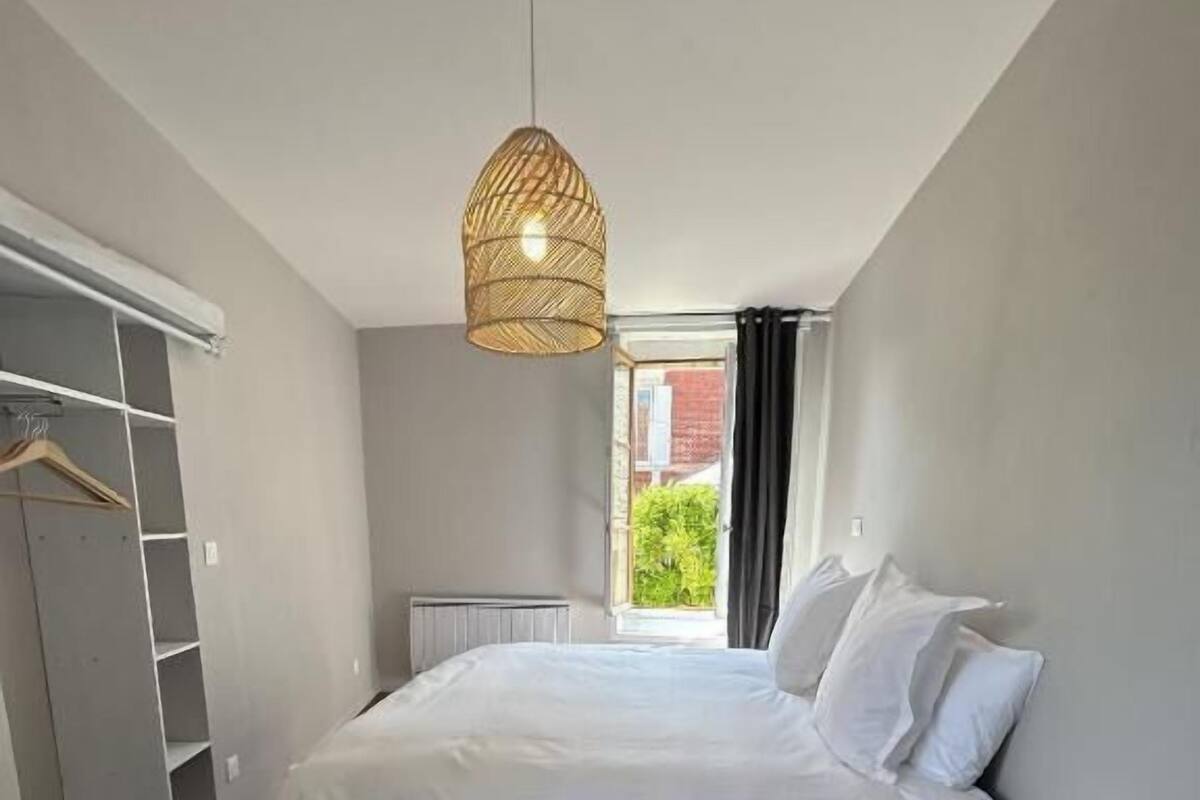 Ein Schlafzimmer mit einem Bett, einer Hängeleuchte, einem Heizkörper, einem Fenster mit Vorhängen und einem weißen Nachttisch mit einem gewobenen Korb.