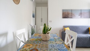 Een eethoek met een tafelkleed met dessin, een potplant, witte stoelen, een grijze bank met gele kussens en een witte kledingkast op de achtergrond.