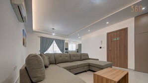 A modern living room with a sectional sofa, a wooden coffee table, and a view of the kitchen and dining area through large windows. - Central Comfort: Stylish 2BR Apt with Balconies! (Qormi)
