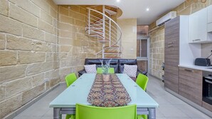 A compact living space with a dining area featuring a long table and green chairs, a built-in sofa, a spiral staircase, and a kitchenette. - Seaview Marsaxlokk Townhouse, steps from Market! (Marsaxlokk)