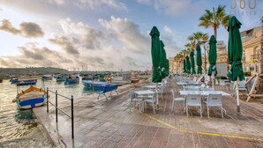 A waterfront café with outdoor seating, boats docked nearby, and a clear sky. - Seaview Marsaxlokk Townhouse, steps from Market! (Marsaxlokk)