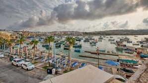 A marina with numerous boats docked, a row of palm trees, and a view of the town in the background. - Seaview Marsaxlokk Townhouse, steps from Market! (Marsaxlokk)