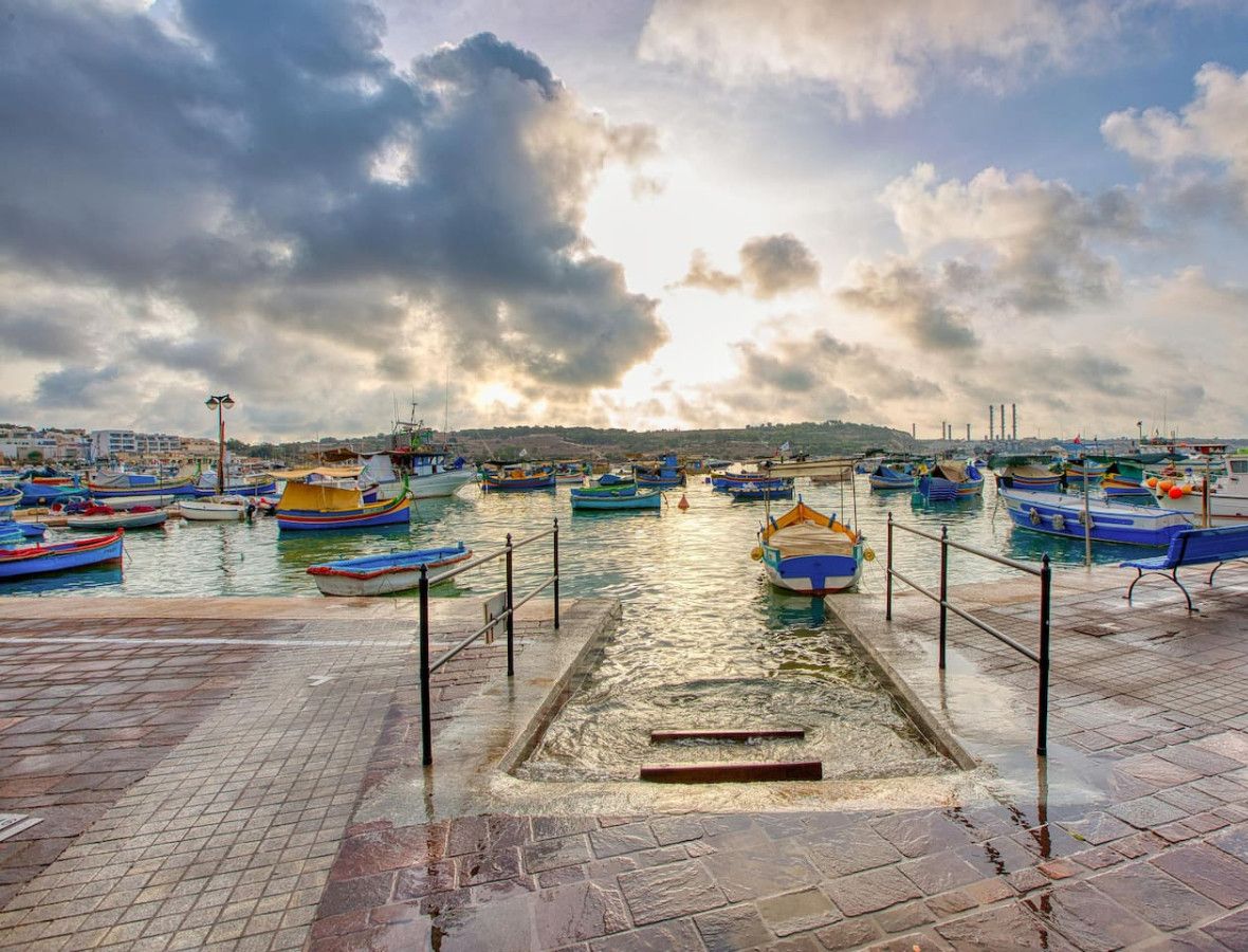 A marina with numerous boats docked, a paved walkway, and a distant view of buildings.