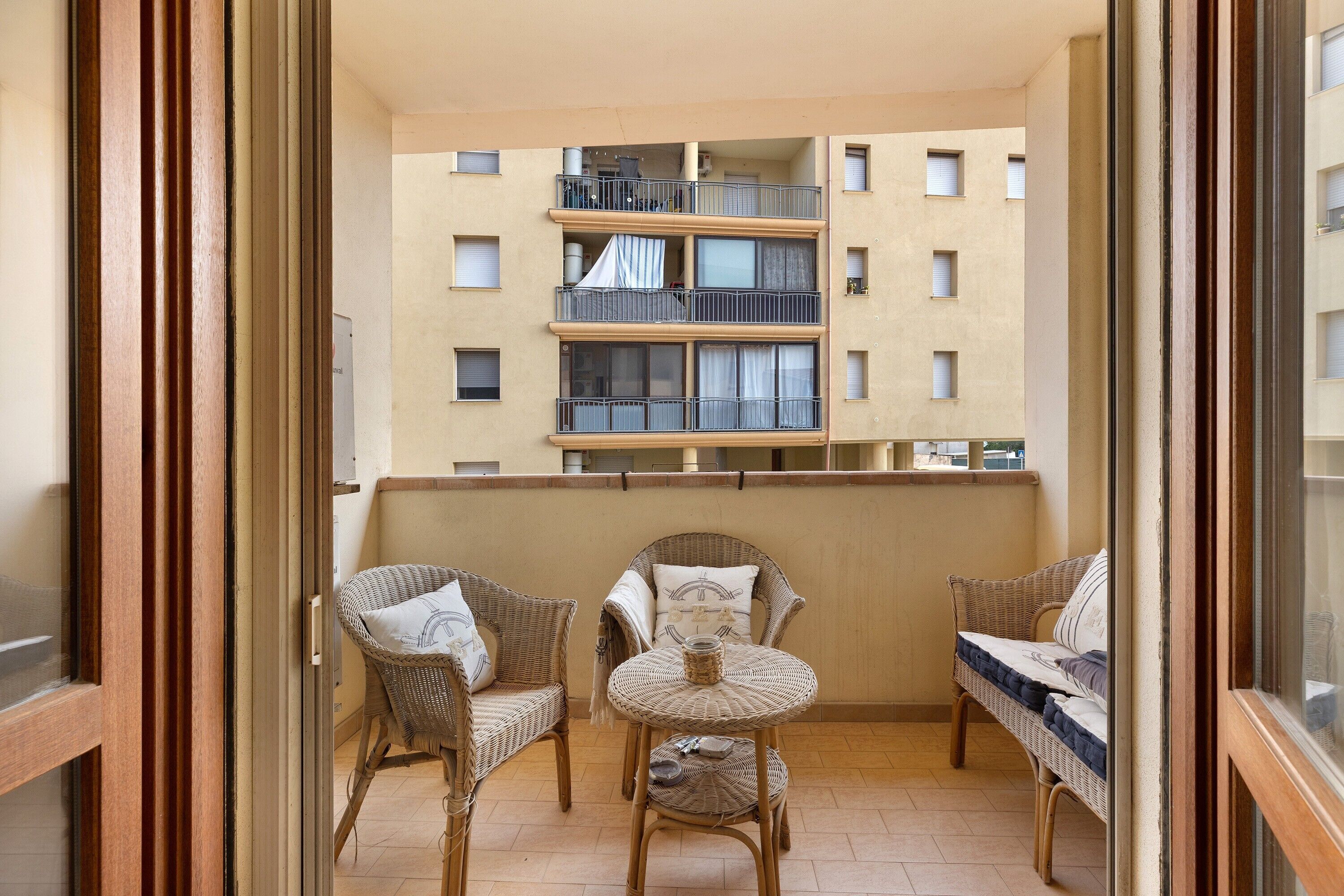 Un balcon doté de meubles en osier, notamment des chaises et une petite table, donnant sur un immeuble comportant de nombreux balcons.