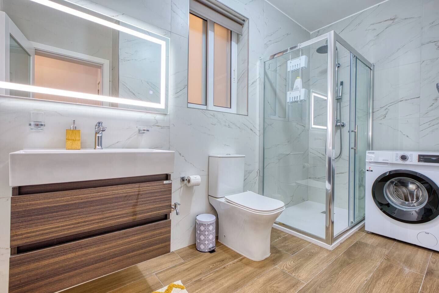 Ein modernes Badezimmer mit einem Holzwaschtisch, einer Toilette, einer Duschkabine aus Glas und einer Waschmaschine mit Frontlader.