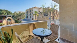 A balcony with wicker chairs and a small round table, overlooking a street with buildings and trees. - Qala Tranquil Escape - Stylish with Terrace & AC (Qala)