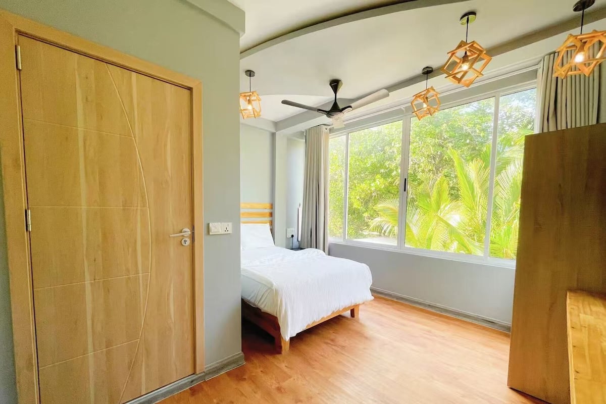 A bedroom with a bed, a wardrobe, a ceiling fan, and large windows with a view of greenery.