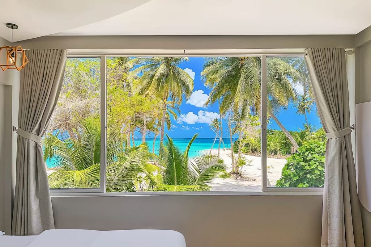 A bedroom with a large window offering a view of a beach and palm trees.