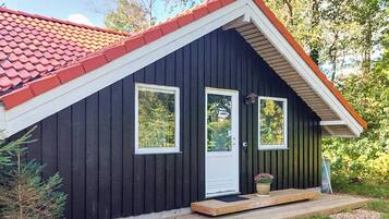 A small, dark wooden cabin with a red-tiled roof, two windows, and a wooden porch.