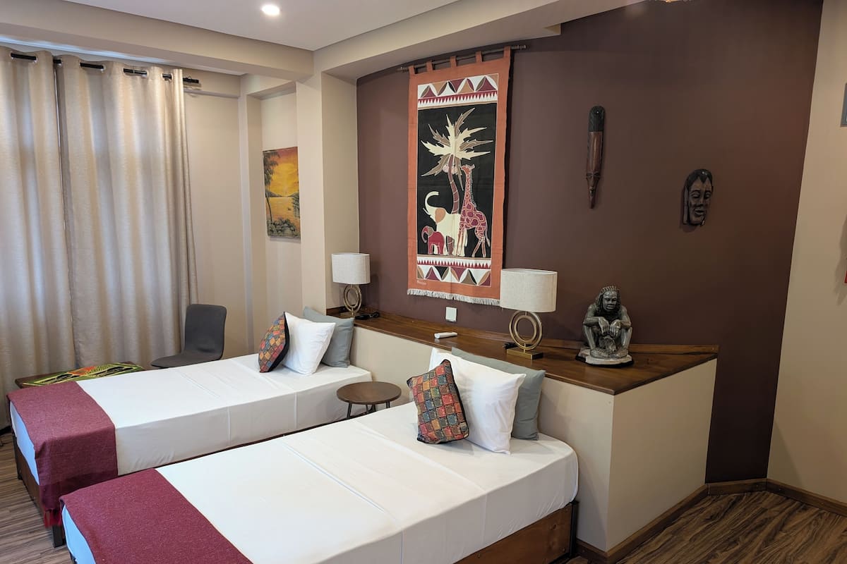 A hotel room with two beds, a desk, a chair, a lamp, and a wall painting.