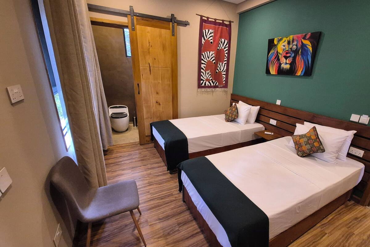 A hotel room with two beds, a chair, a wooden door, a curtain, a painting, and a trash bin.