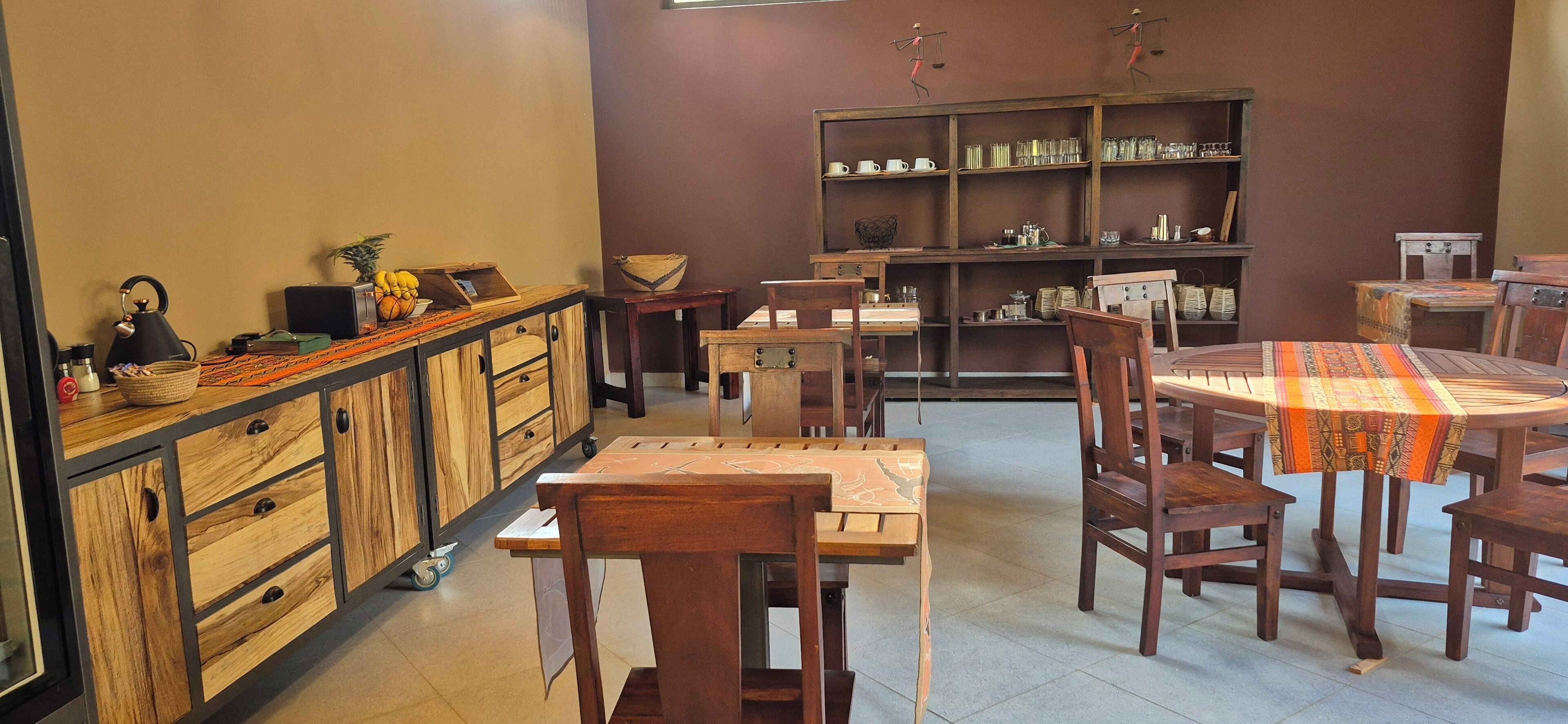 Una habitación acogedora y bien iluminada con muebles de madera, que incluyen una mesa de comedor con sillas, una mesita auxiliar y un aparador con cajones. Hay una estantería con varios objetos, y una escultura decorativa en la pared.
