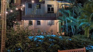 Un edificio de dos pisos con un balcón, rodeado de abundante vegetación y luces colgantes.