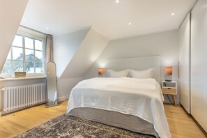Ein modernes Schlafzimmer mit einem großen Bett, zwei Nachttischen mit Lampen, einem Heizkörper und Blick auf ein Haus durch ein Fenster.