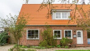 Une maison à deux étages, construite en briques, avec un toit recouvert de tuiles rouges, deux fenêtres au rez-de-chaussée et un petit porche.