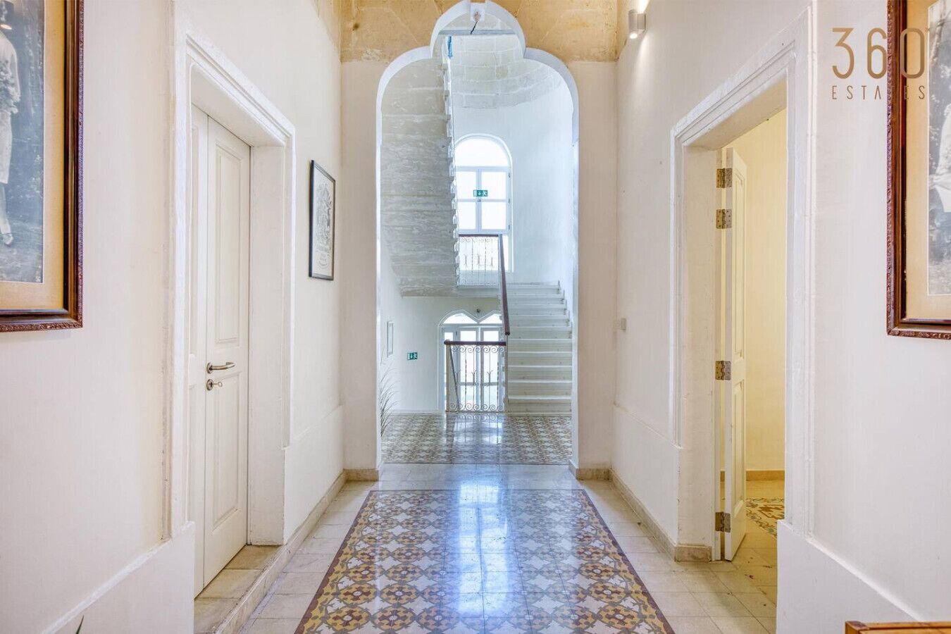 A corridor with a tiled floor, white walls, and a staircase leading to a balcony.