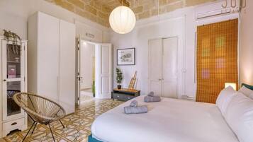 A bedroom with a large bed, a wicker chair, a door leading to another room, and a wall-mounted light fixture.