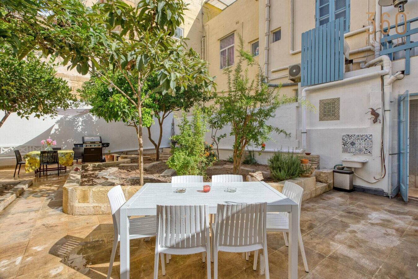 An outdoor patio with a white table and chairs, surrounded by greenery and a stone floor.