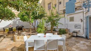 An outdoor patio with a white table and chairs, surrounded by greenery and a stone floor.