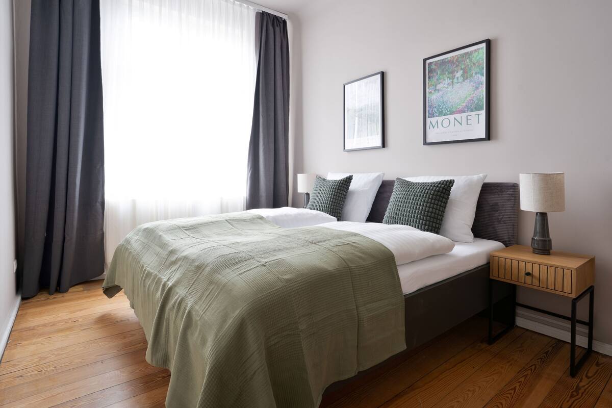 A bedroom with a bed, a nightstand, a lamp, a framed picture, and a window with curtains.