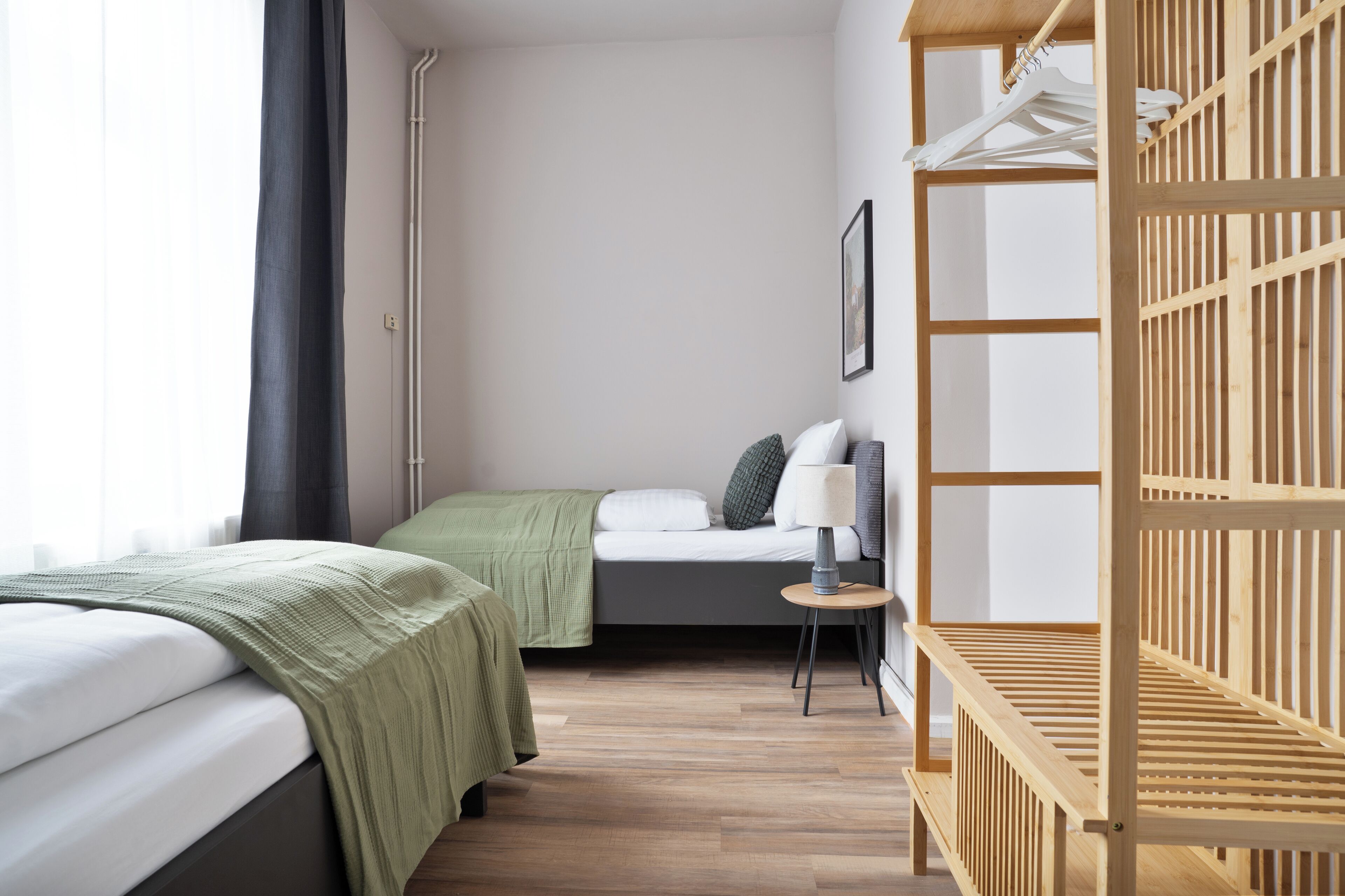 A hotel room with two beds, a wooden shelf unit, a small table with a lamp, and a large window with curtains.