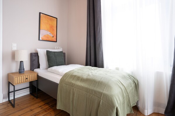 A neatly arranged bedroom with a bed, a bedside table with a lamp, a wall-mounted painting, and a window with curtains. - Schöne Apartments in Dessau I home2share (Dessau-Roßlau)