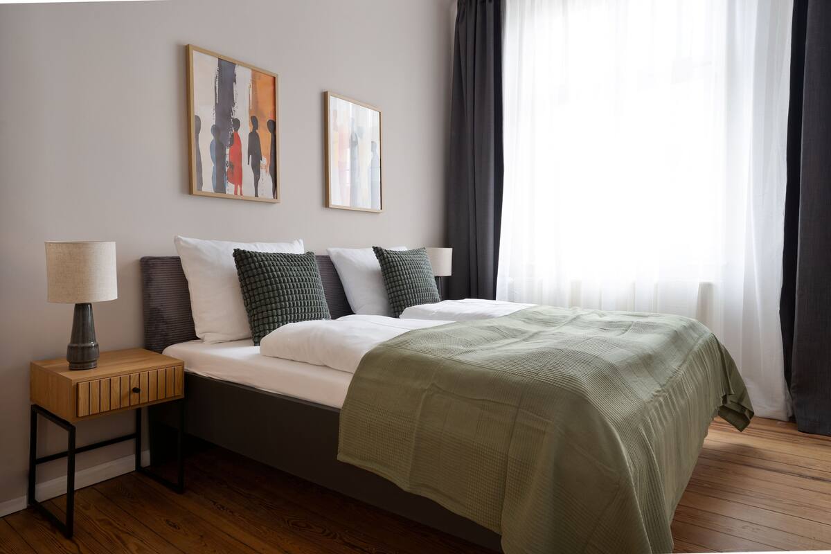 A neatly arranged bedroom with a bed, a bedside table with a lamp, and two framed artworks on the wall.