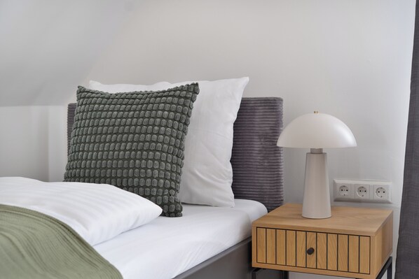 Una cama bien hecha con un cojín verde y blanco, un cojín gris y una lámpara blanca sobre una mesita de noche de madera con un enchufe.