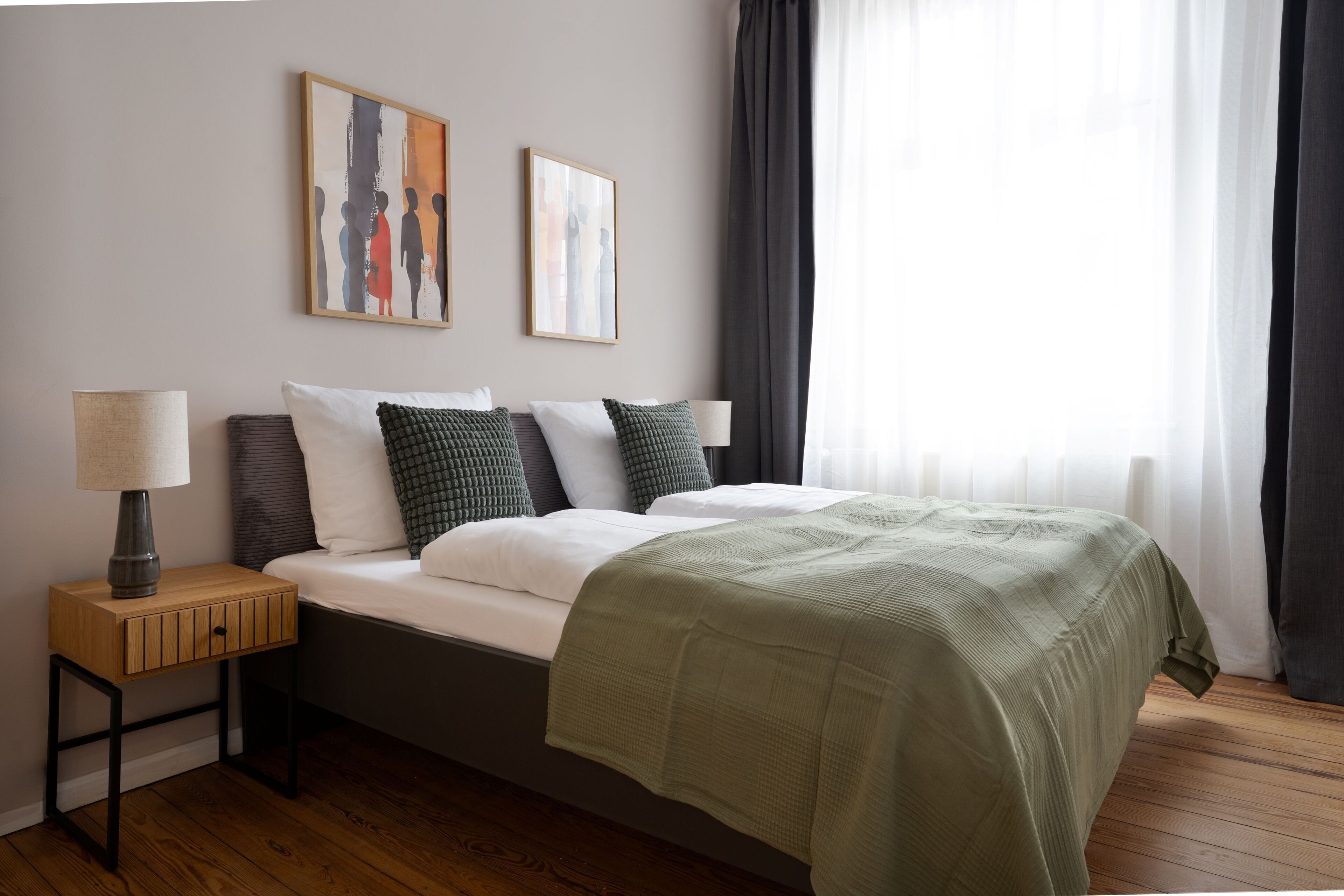 Een keurig opgemaakt slaapkamer met een bed, een nachtkastje met een lamp, en twee ingelijste kunstwerken aan de muur.
