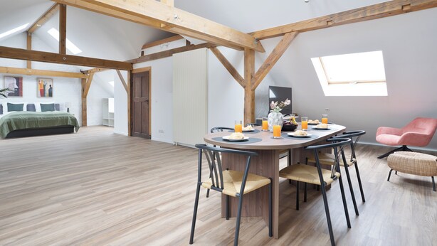 Ein geräumiges Loft mit hohen Decken, freiliegenden Holzbalken, einem Essbereich mit Tisch und Stühlen sowie einem Bett im Hintergrund.