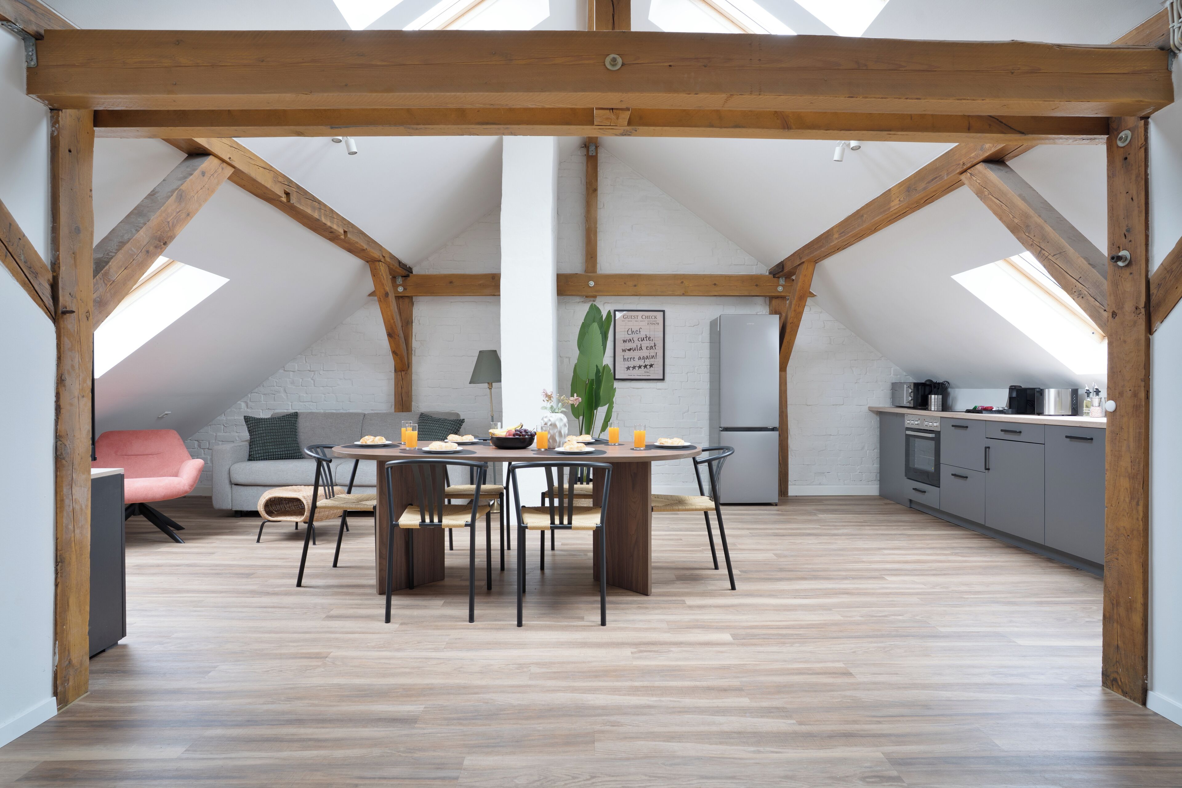 Un loft spacieux avec de hauts plafonds, des poutres apparentes, un coin repas avec une table ronde et des chaises, une kitchenette et une grande fenêtre qui laisse entrer la lumière naturelle.