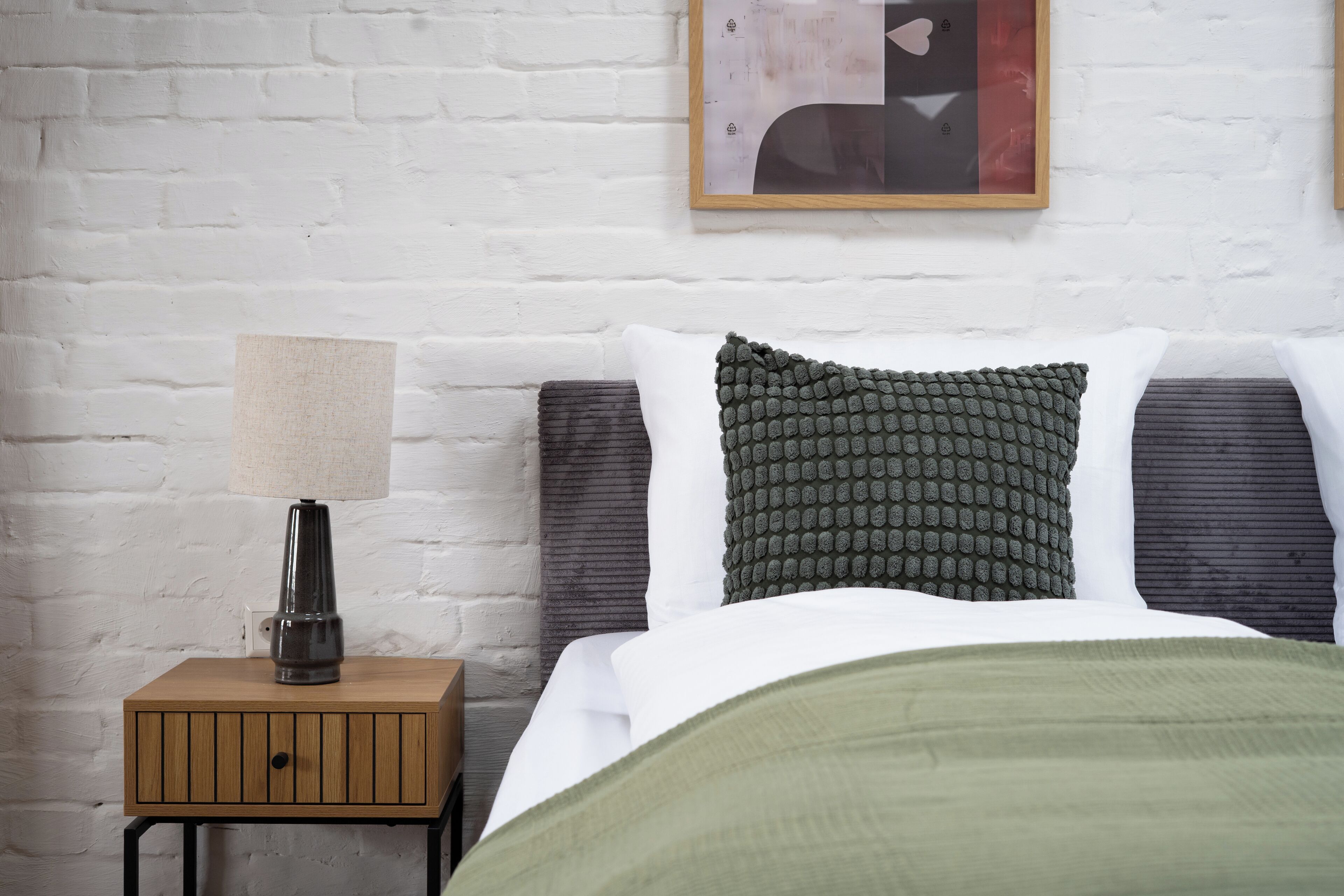 Una cama bien tendida con una manta verde, una mesita de noche de madera con una lámpara y una pared de ladrillo blanco.